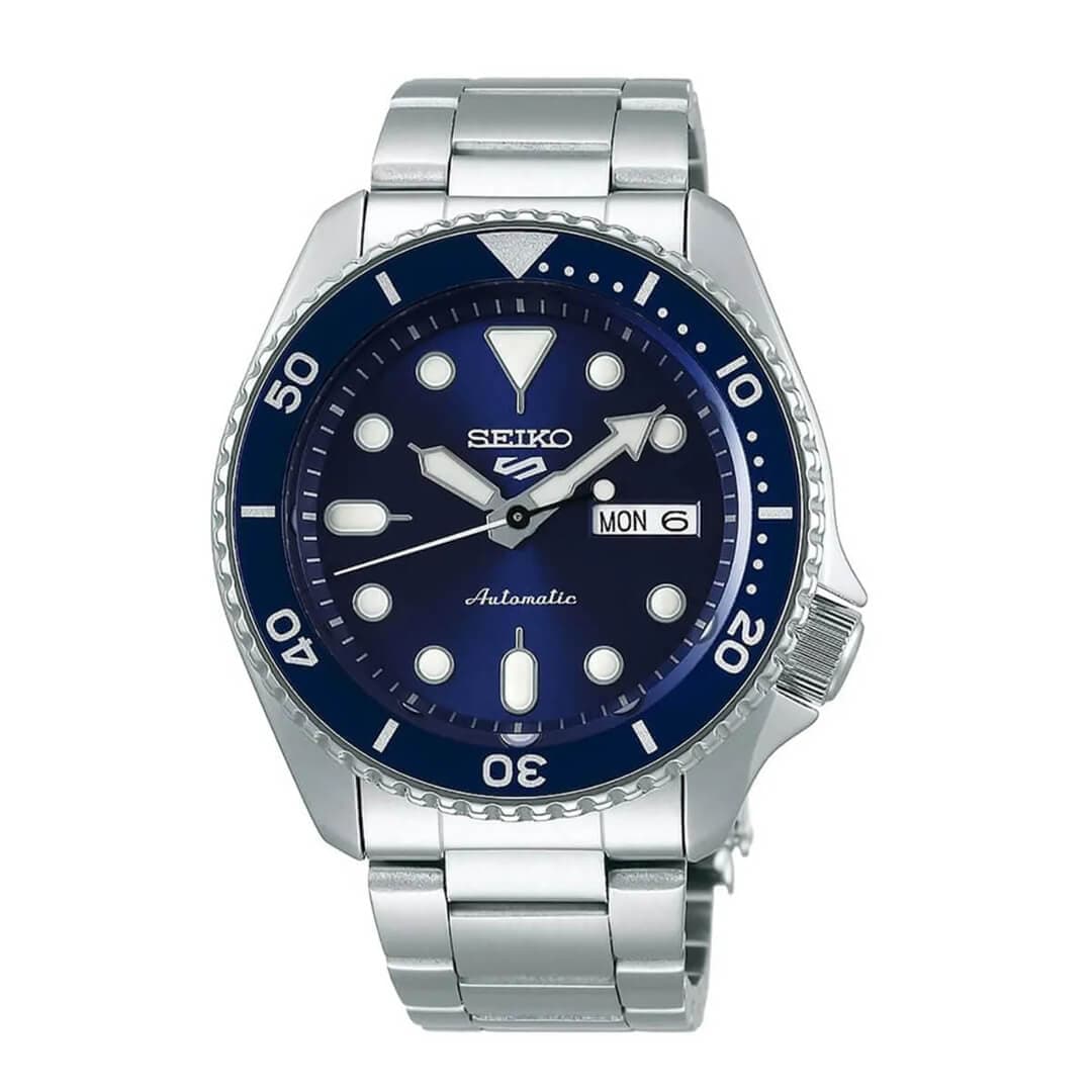 Sølvfarget Seiko automatisk klokke med marineblå urskive, selvlysende visere, sirkulære timemarkører og datovisning.